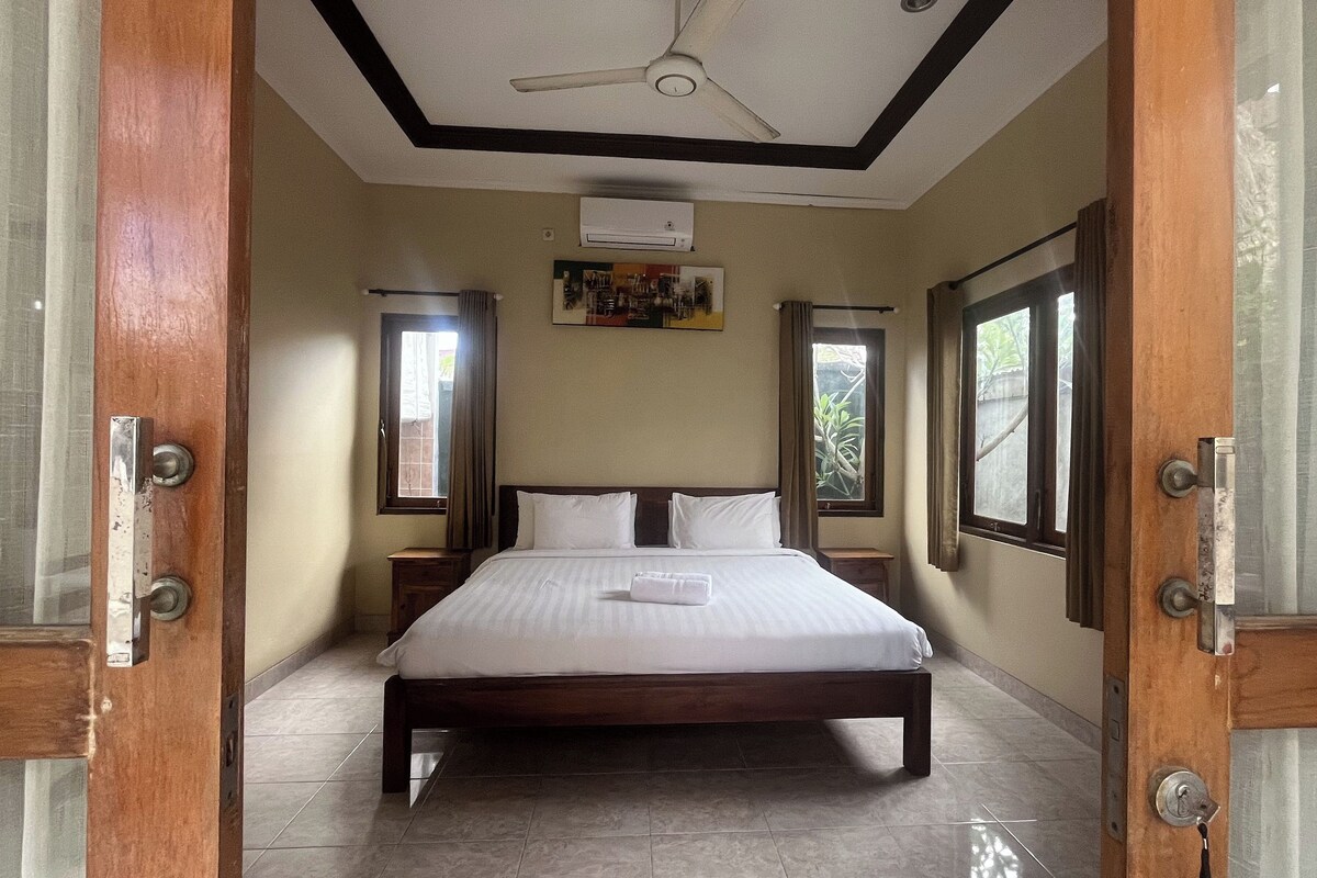 A hotel room with a large bed, wooden headboard, two bedside tables, an air conditioning unit, and large windows with curtains.
