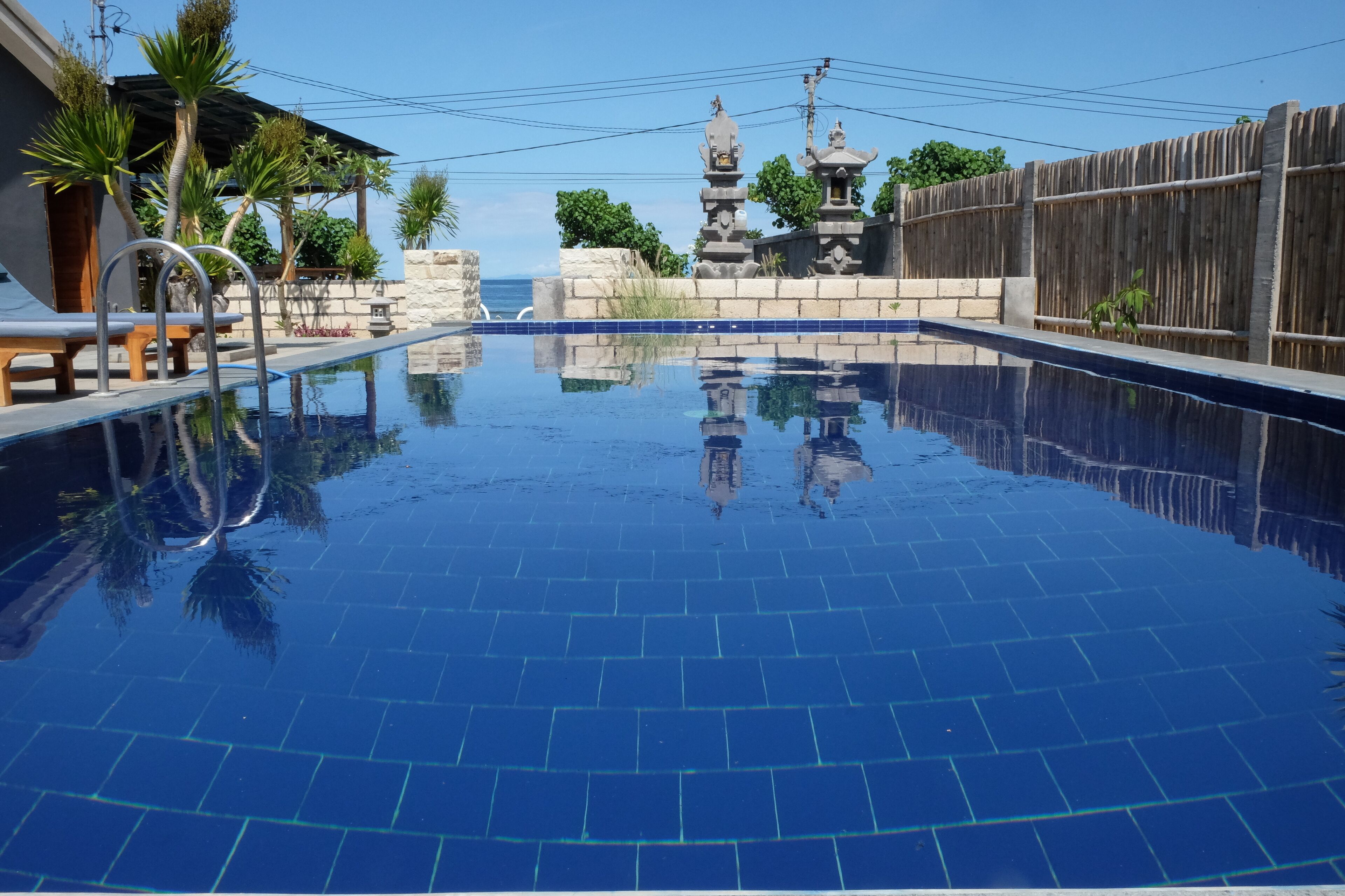 A swimming pool with clear blue water, surrounded by a wooden fence, stone walls, and a small pagoda-like structure.
