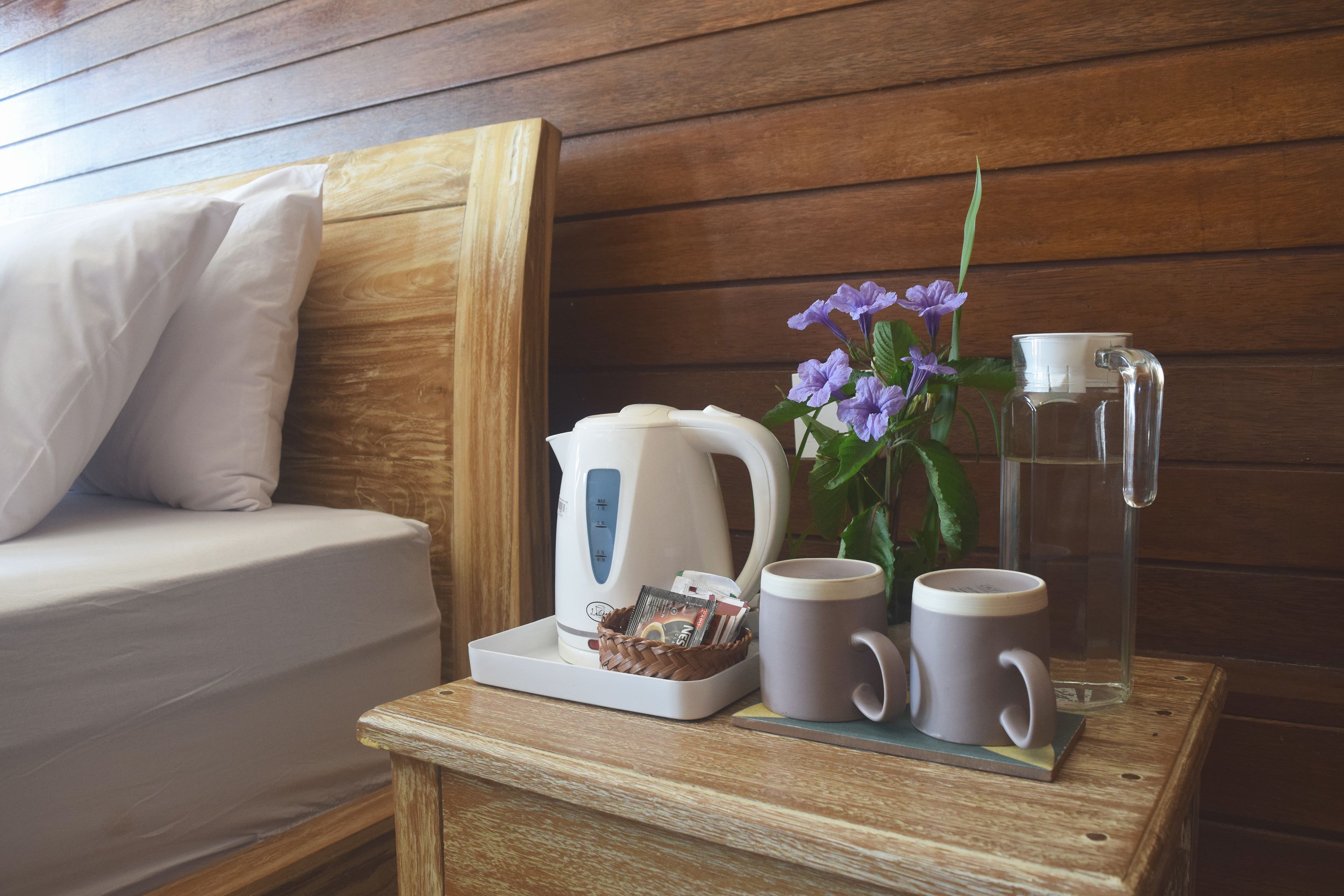 A wooden bedside table with a white kettle, two mugs, a glass pitcher, and a basket of magazines.