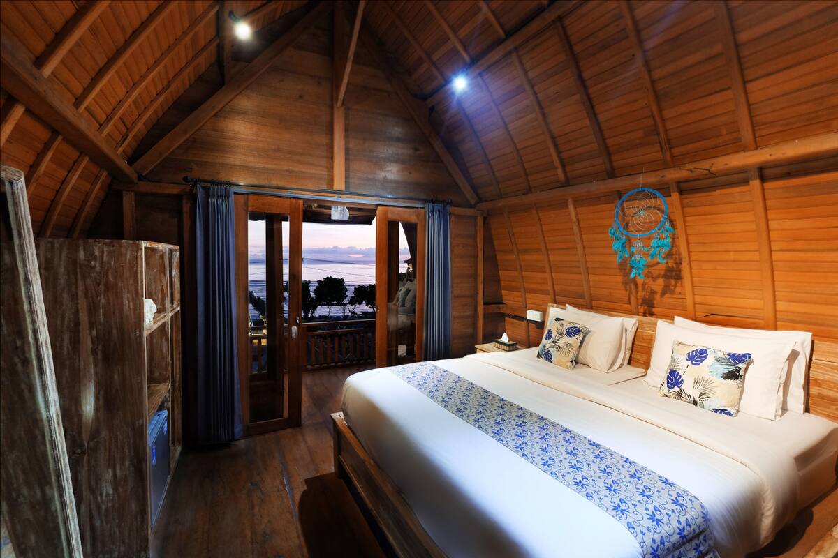 A cozy, wooden-floored bedroom with a large bed, a dreamcatcher, and a view through an open door.