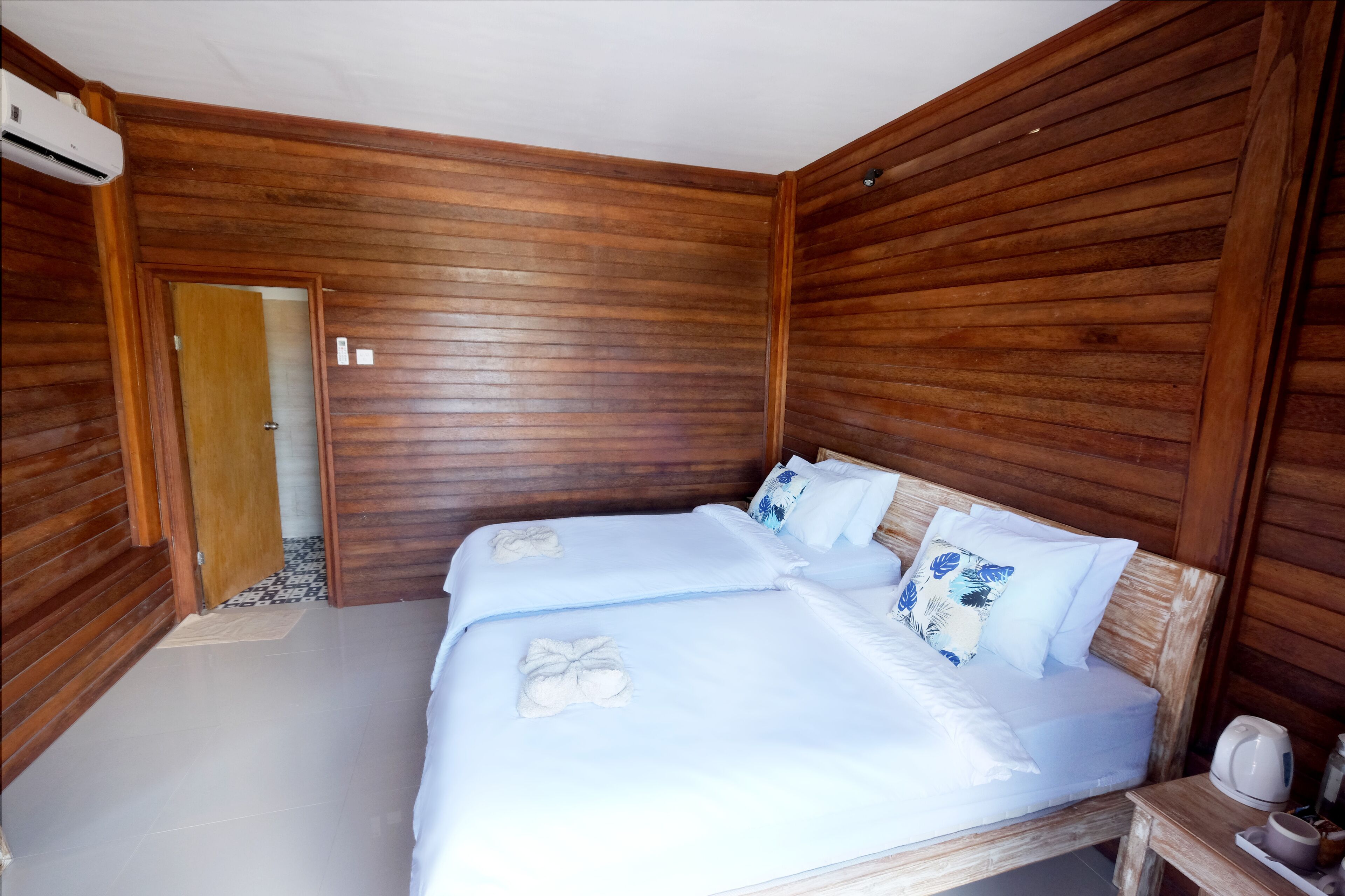 A room with wooden paneling, a bed with white linens and decorative pillows, a bedside table with a kettle, and an air conditioning unit.