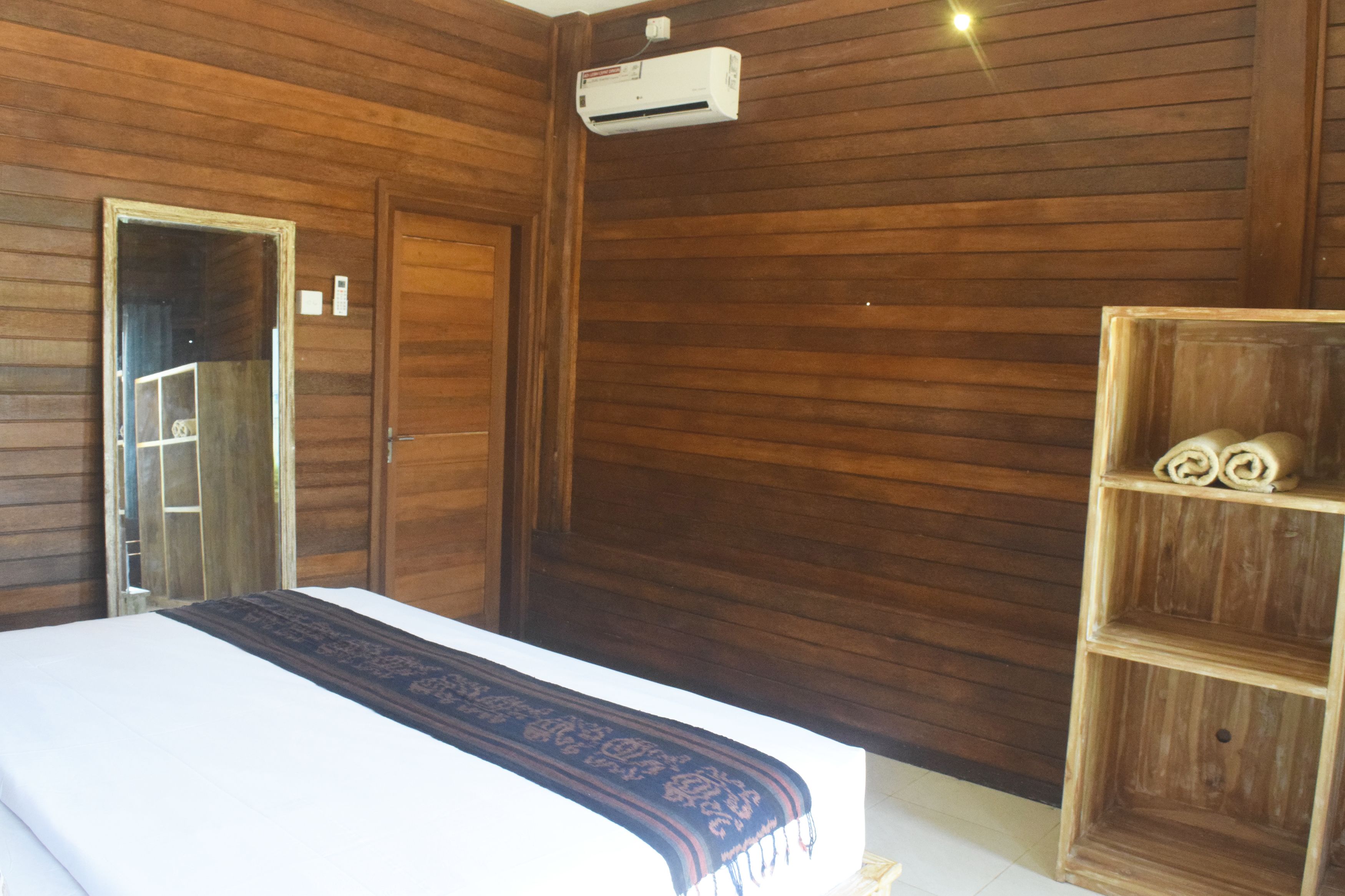 A wooden-paneled room with a bed, a mirror, an air conditioning unit, and a wooden shelf with rolled towels.