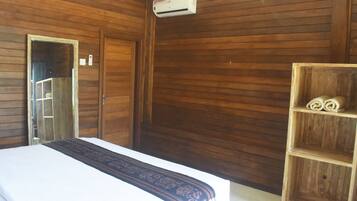Habitación con paneles de madera, una cama, un espejo, un equipo de aire acondicionado y un estante de madera con toallas enrolladas.
