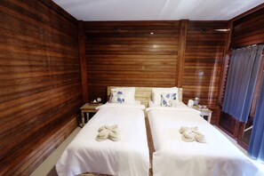 A room with two beds, wooden paneling, a nightstand with a lamp, and a window with curtains.