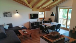 Una sala de estar moderna con un sofá, una mesa de centro y un comedor. Un televisor está montado en la pared, y hay una ventana con vistas a zonas verdes.