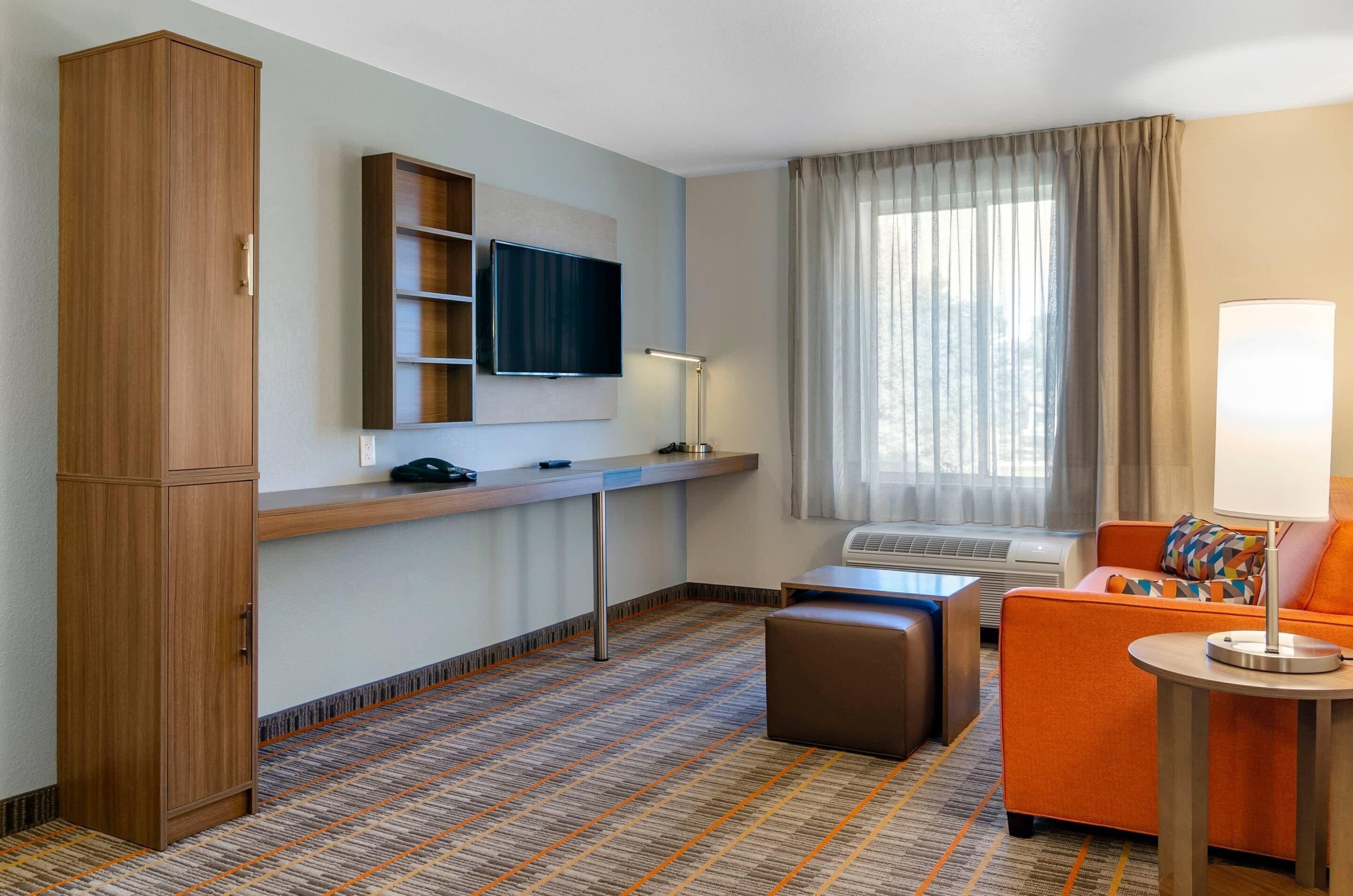 Quarto de hotel com televisão, sofá, apoio para os pés e secretária com luminária.
