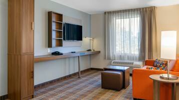 Quarto de hotel com televisão, sofá, apoio para os pés e secretária com luminária.