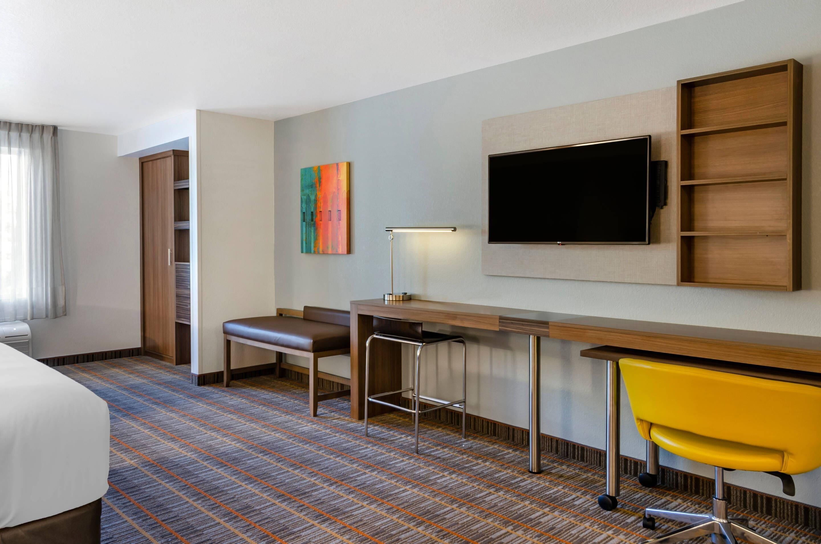 A hotel room with a bed, a desk with a chair, a TV, a bench, and a colorful painting.