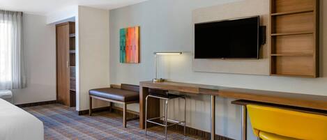 A hotel room with a bed, a desk with a chair, a TV, a bench, and a colorful painting.