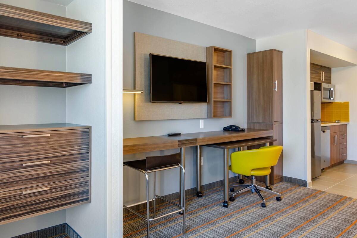 A modern hotel room with a wooden desk, a flat-screen TV, a yellow office chair, and a kitchen area.