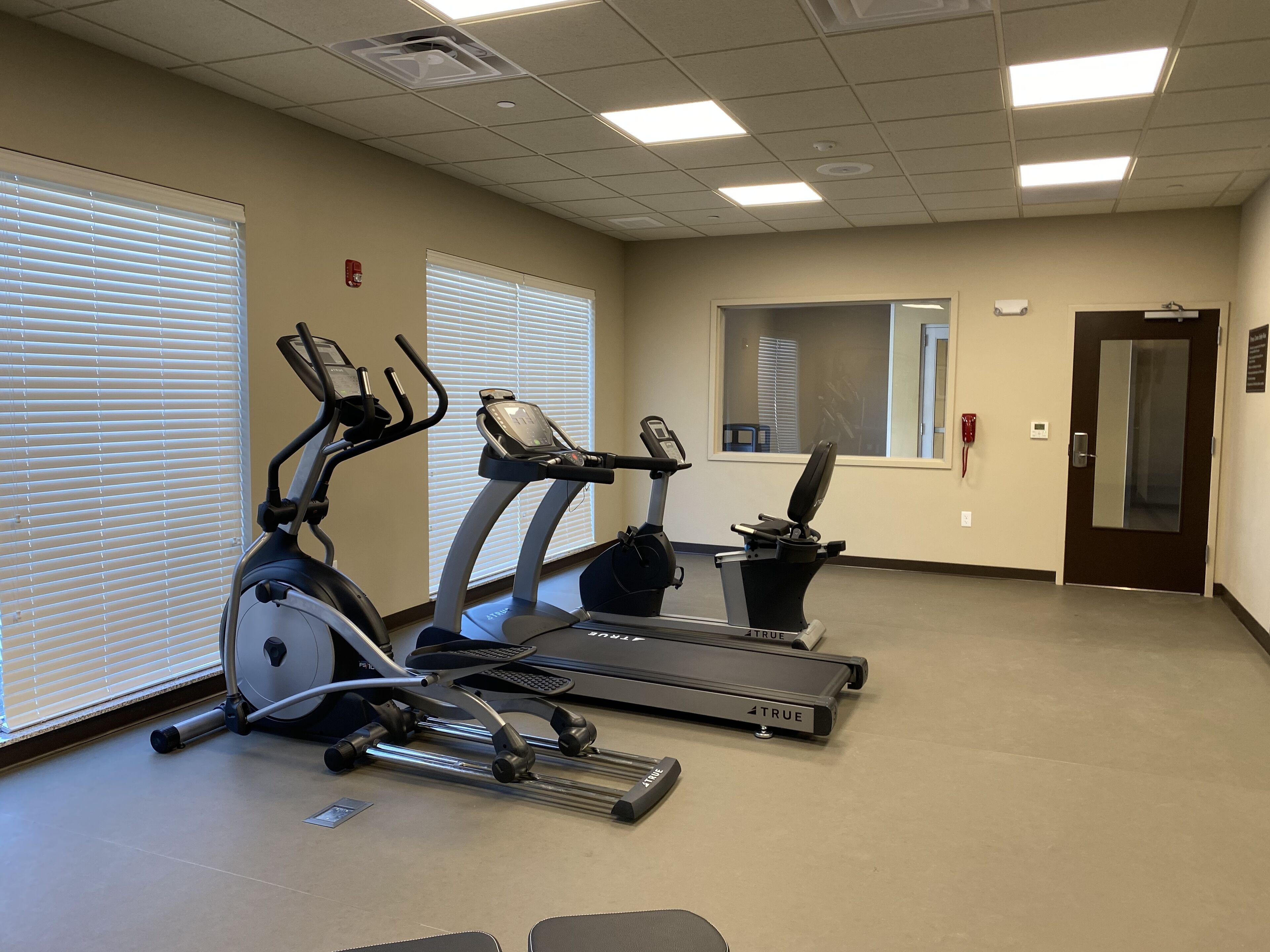 A fitness room with three treadmills, a mirrored wall, and a door with a red emergency phone.