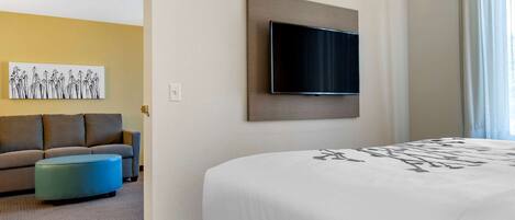 Ein Hotelzimmer mit Bett, einem an der Wand befestigten Fernseher, einer Couch und einem blauen Ottomanen.