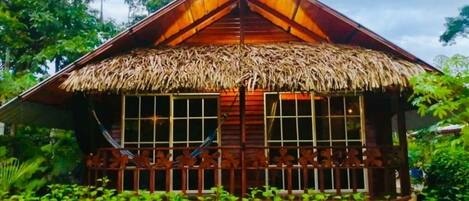 A wooden house with a thatched roof, surrounded by lush greenery.