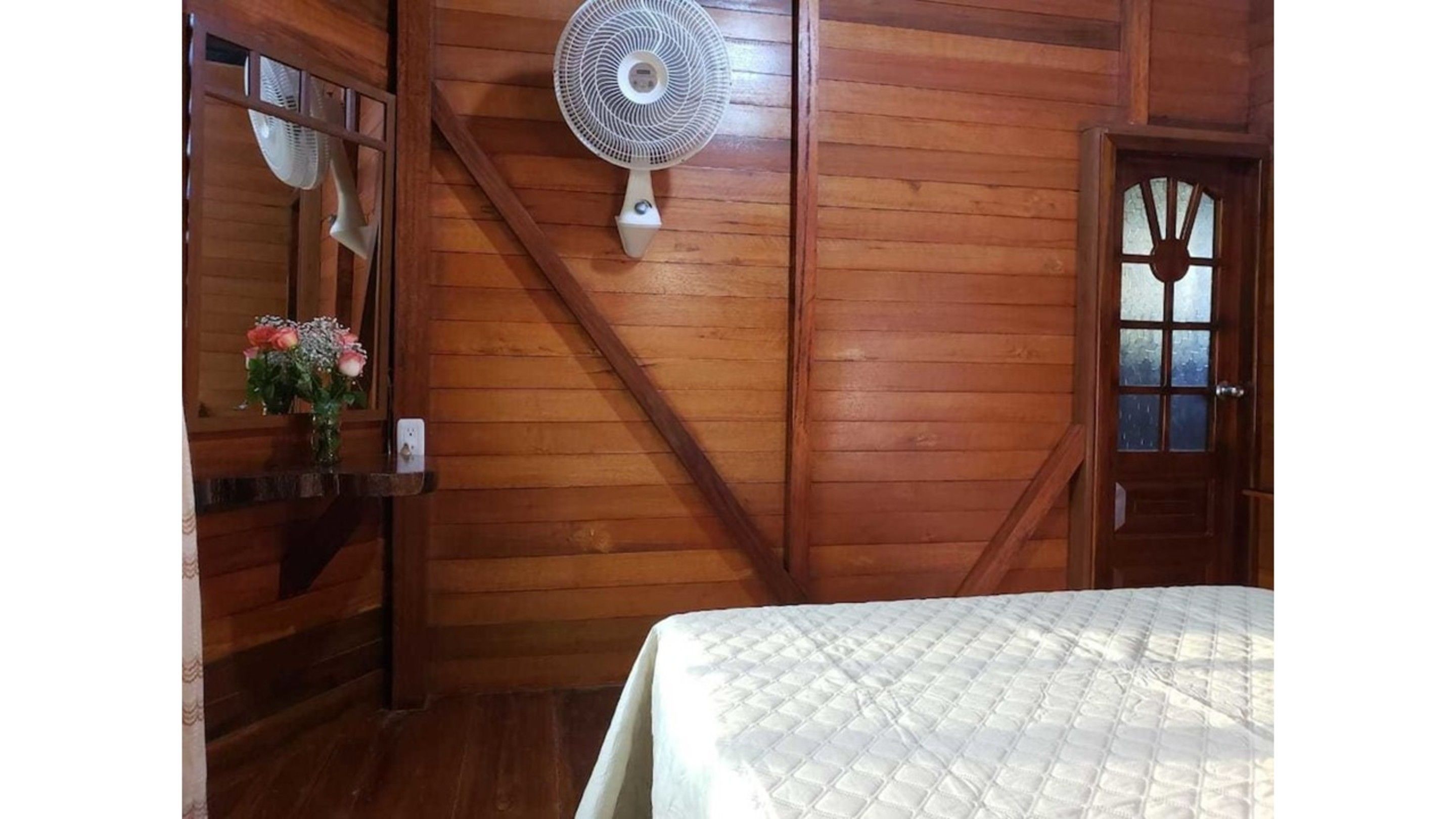 A wooden room with a bed, a wall-mounted fan, and a door with a window.