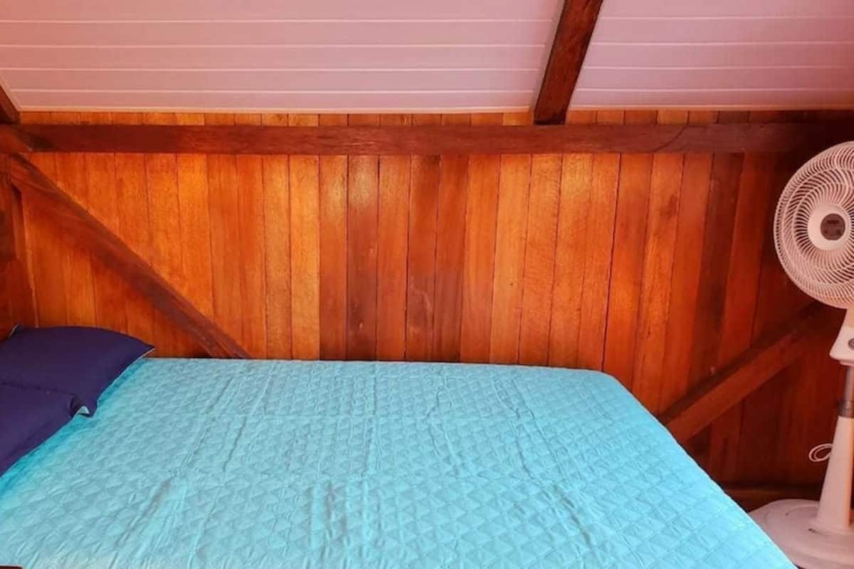 A small, cozy room with a wooden ceiling and walls, a single bed with a blue quilt, and a standing fan.