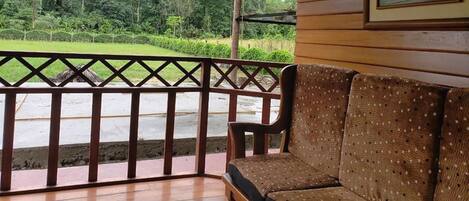 Una terraza de madera con un sofá tejido y una sola silla, con vistas a un paisaje verde con una valla.