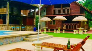 Una terraza de madera con sillones de descanso y una sombrilla azul, una piscina y una casa de madera con sombrillas de paja al fondo.