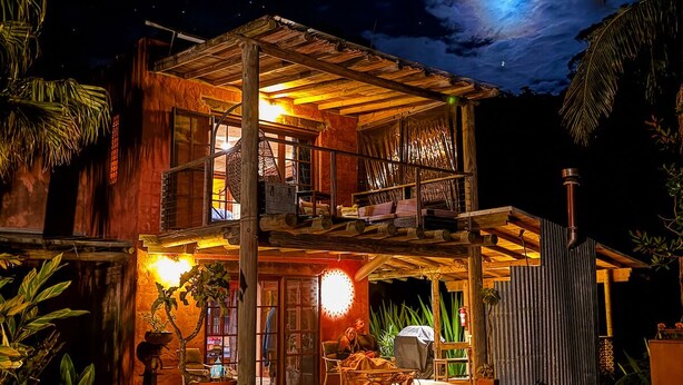 Una casa de fusta de dos pisos amb un balcó, envoltada de plantes tropicals i un cel nocturn amb la lluna visible.