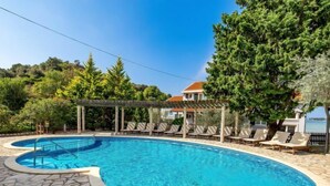 A swimming pool with lounge chairs and a pergola, surrounded by greenery and a building in the background.