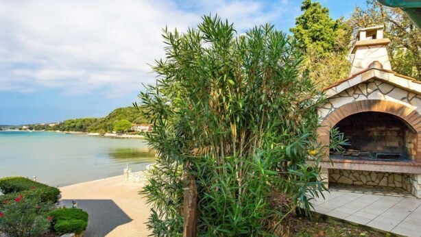 A stone-built outdoor kitchen with a view of a calm lake and a paved pathway leading to a beach.