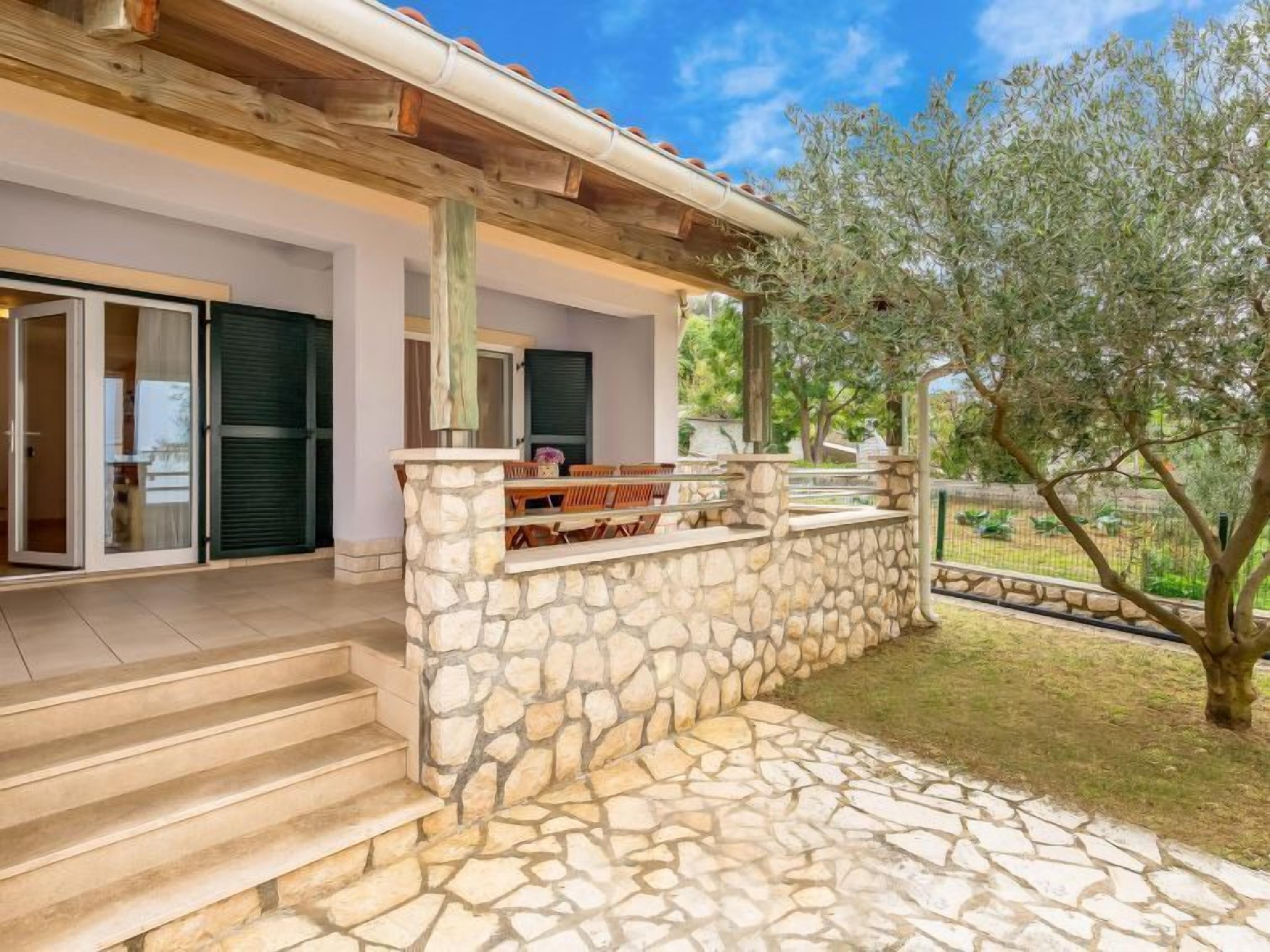 A stone-built outdoor area with a wooden pergola, steps leading to a covered seating area, and a view of a garden with trees.