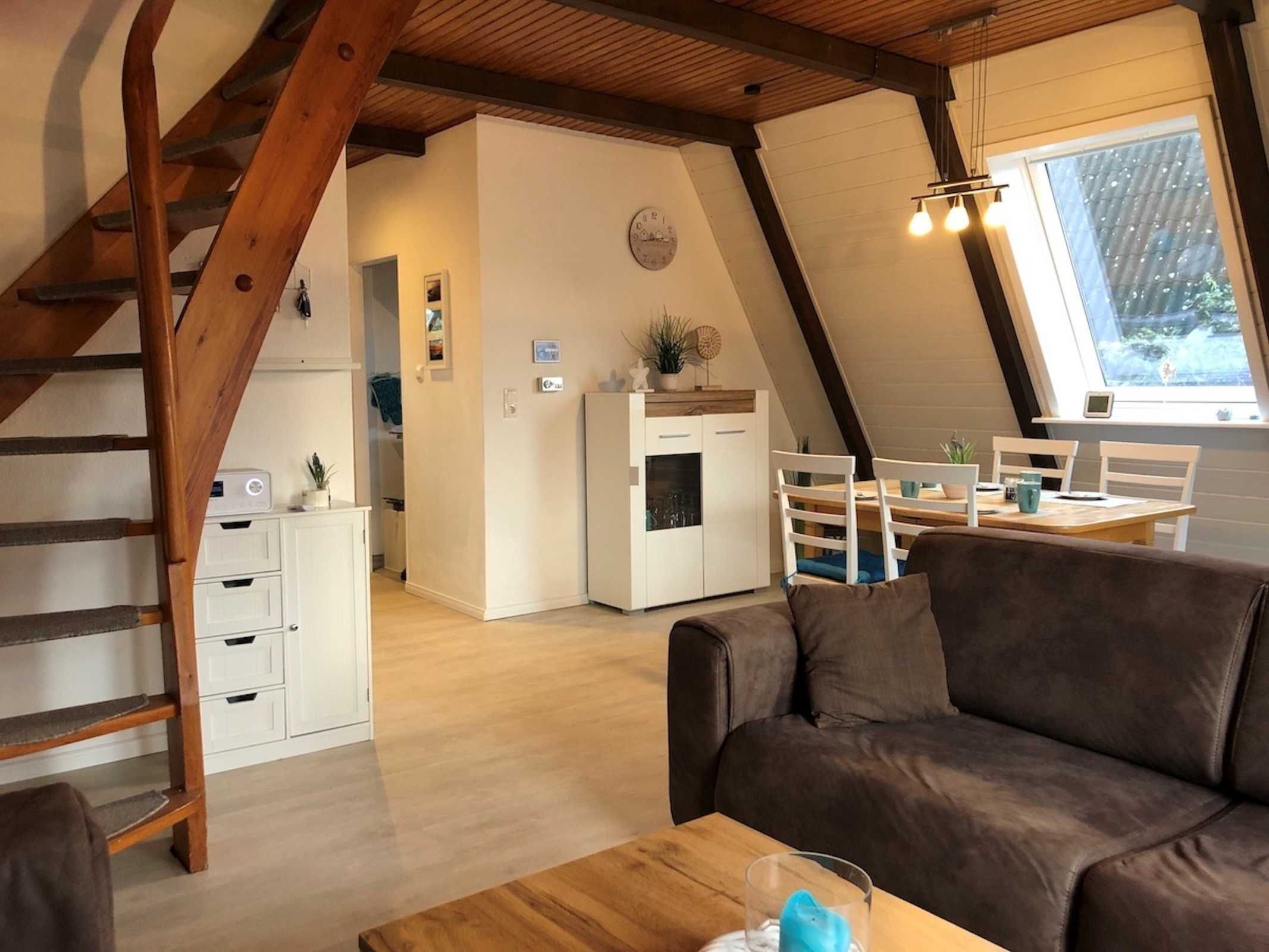 A cozy loft-style living space with a wooden staircase, a kitchenette, a dining area, and a large window providing natural light.