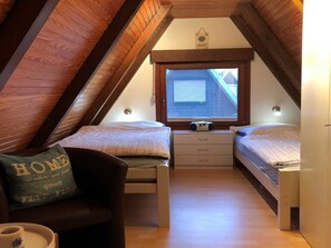 A small, cozy attic bedroom with two beds, a window, and a wooden ceiling.