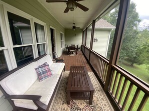 A covered porch with a wooden bench, a white sofa with a plaid pillow, and a view of a green landscape.
