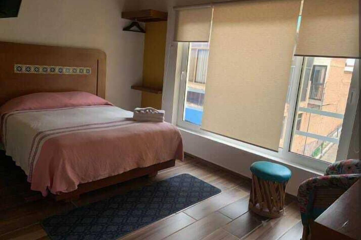 Un dormitorio con una cama, una ventana con persianas, un taburete pequeño y suelo de madera.