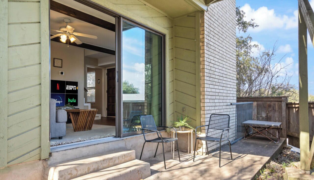 Un patio con mobiliario exterior, un ventilador de techo y un gran ventanal que da a una habitación con televisor y planta.