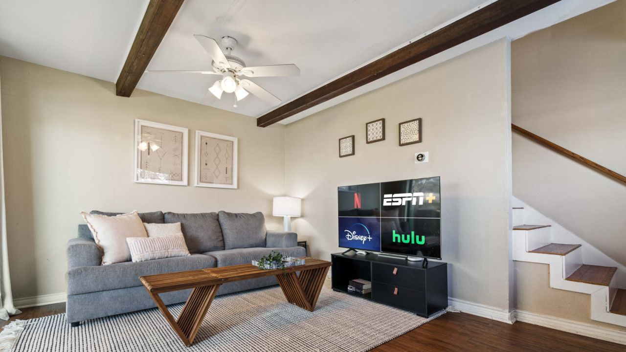 Un soggiorno con un divano grigio, un tavolino in legno e una TV che mostra ESPN, Disney+ e Hulu. Un ventilatore a soffitto e quadri incorniciati adornano la parete.