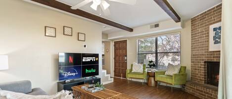 Un salón con televisión mostrando los canales ESPN, Disney+ y Hulu, chimenea, ventilador de techo y pared de ladrillo.