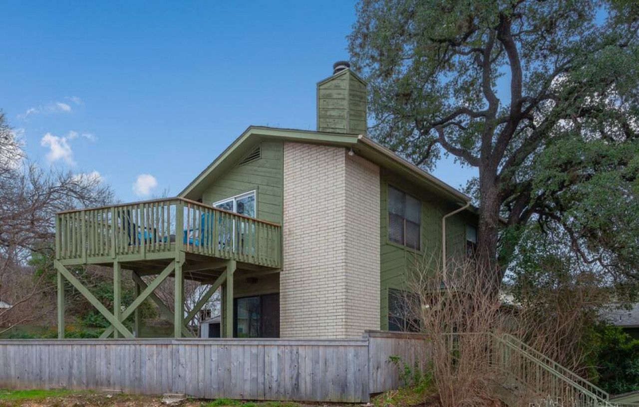 Una casa de dos pisos con fachada verde, una chimenea de ladrillo y una terraza de madera.