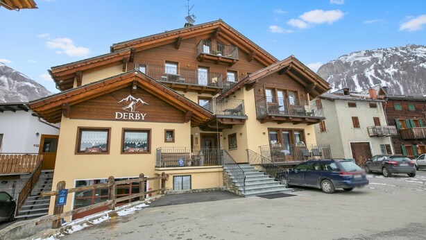 Un immeuble à plusieurs étages, sur lequel on peut lire l’inscription « DERBY », doté de balcons, d’un escalier et d’un espace de stationnement.