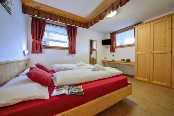 A wooden bed with red bedding, a wooden desk, a television, and a wardrobe in a room with wooden floors and white walls. - Tamigi, Livigno, Italy (Livigno)