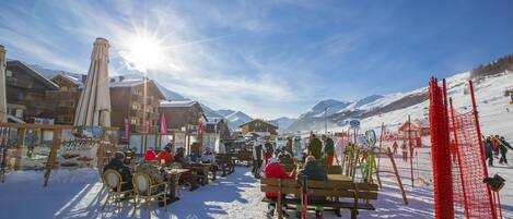 A snow-covered outdoor seating area with wooden benches and tables, surrounded by buildings and a ski slope in the background.