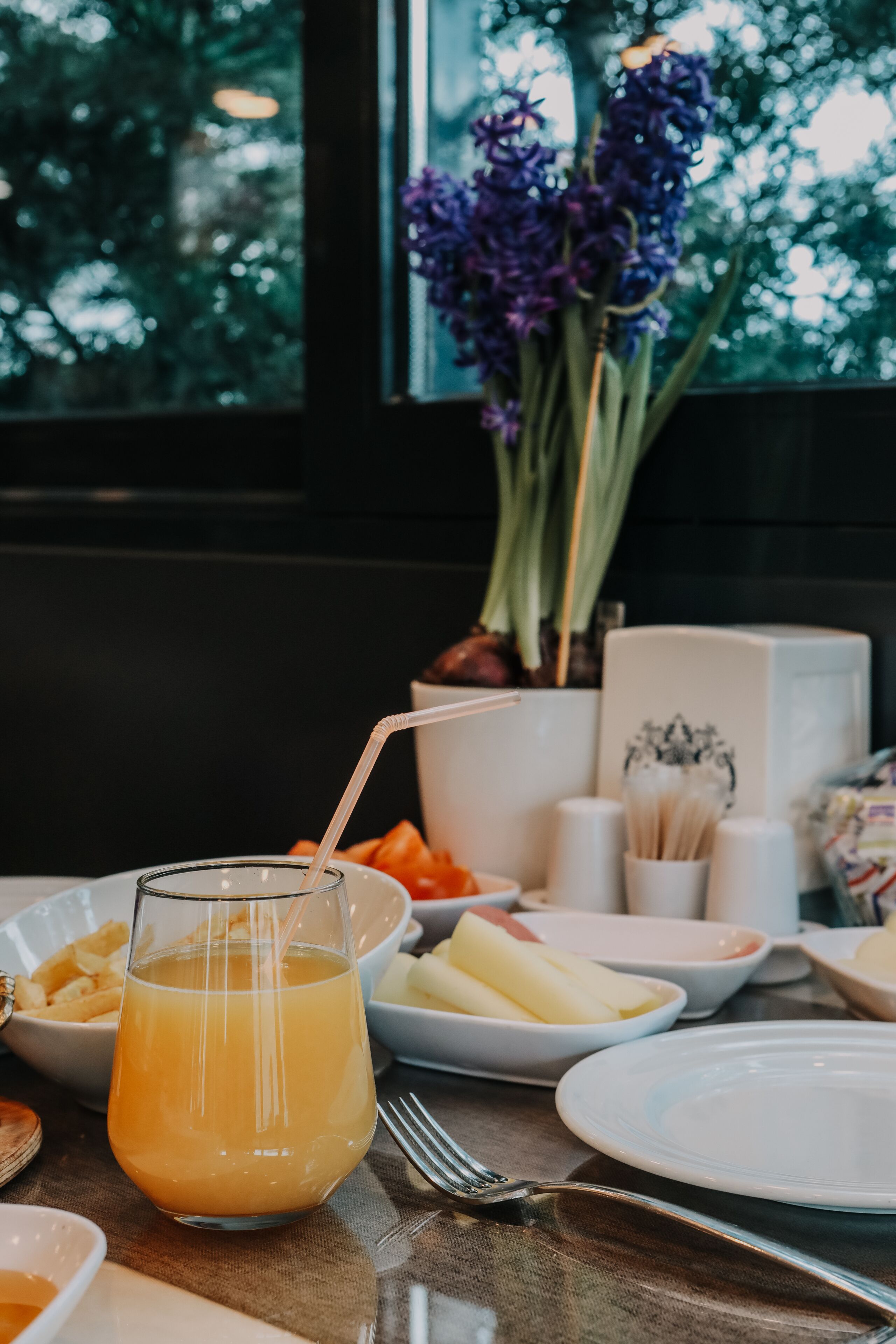 Une table avec un verre de jus d’orange, des assiettes remplies de nourriture et un vase de fleurs violettes.