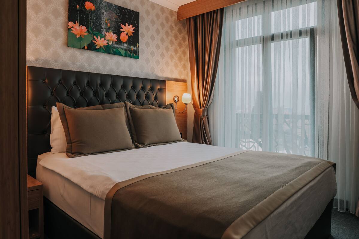 A hotel room with a large bed, a headboard, a nightstand with a lamp, a painting on the wall, and a window with curtains.