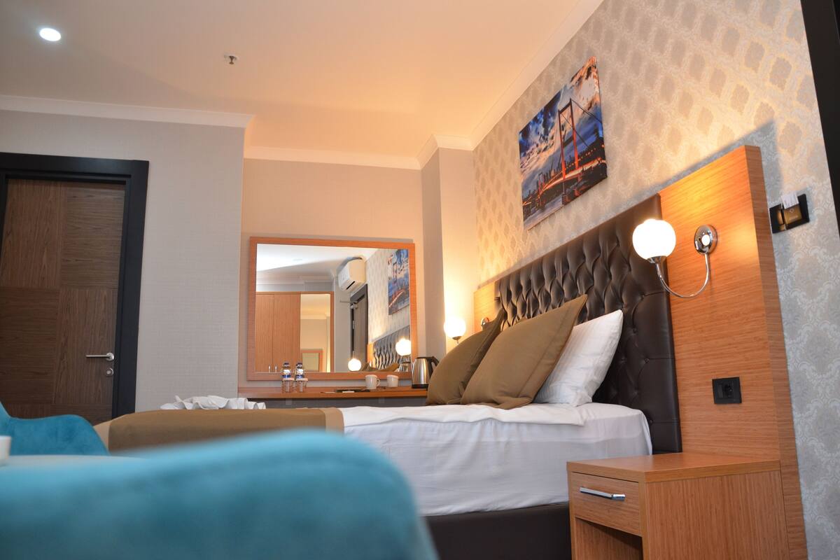 A hotel room with a large bed, a wooden nightstand, a mirror, and a wall-mounted light fixture.