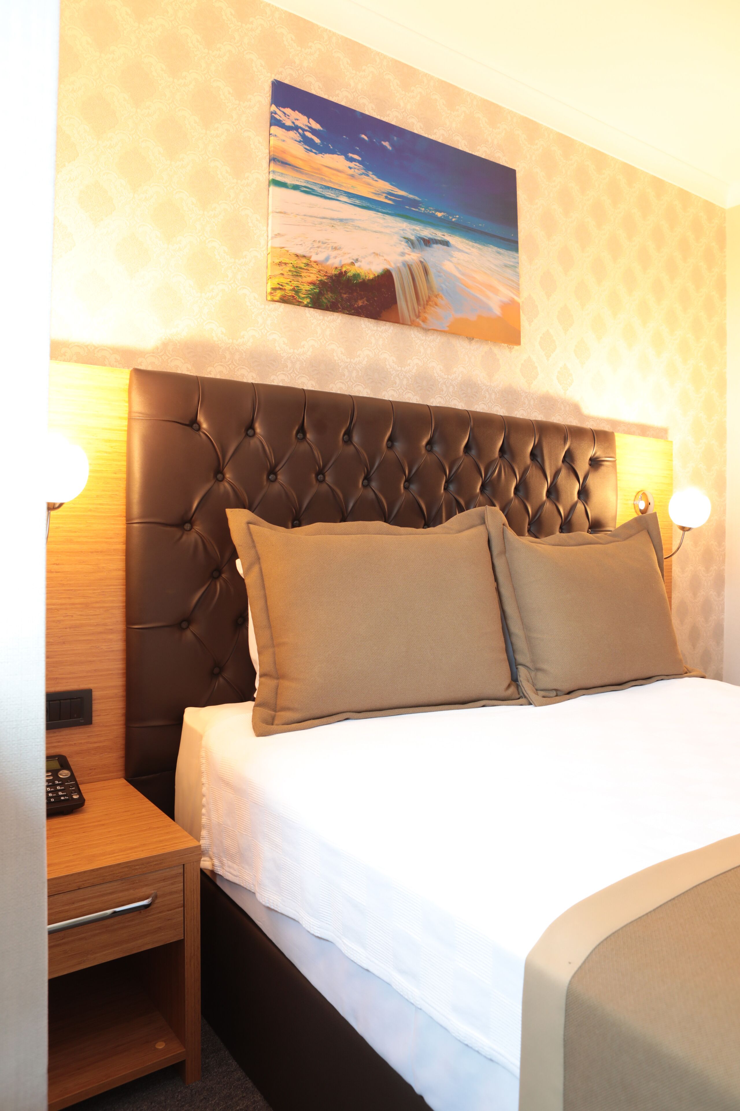 Una habitación de hotel con un cabecero capitonado, un cuadro de una escena de playa, una mesita de noche con una lámpara y otro pequeño mueble con un teléfono.