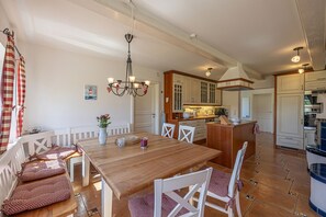 Un comedor con una mesa de madera y sillas blancas, una lámpara de araña, y una cocina al fondo.