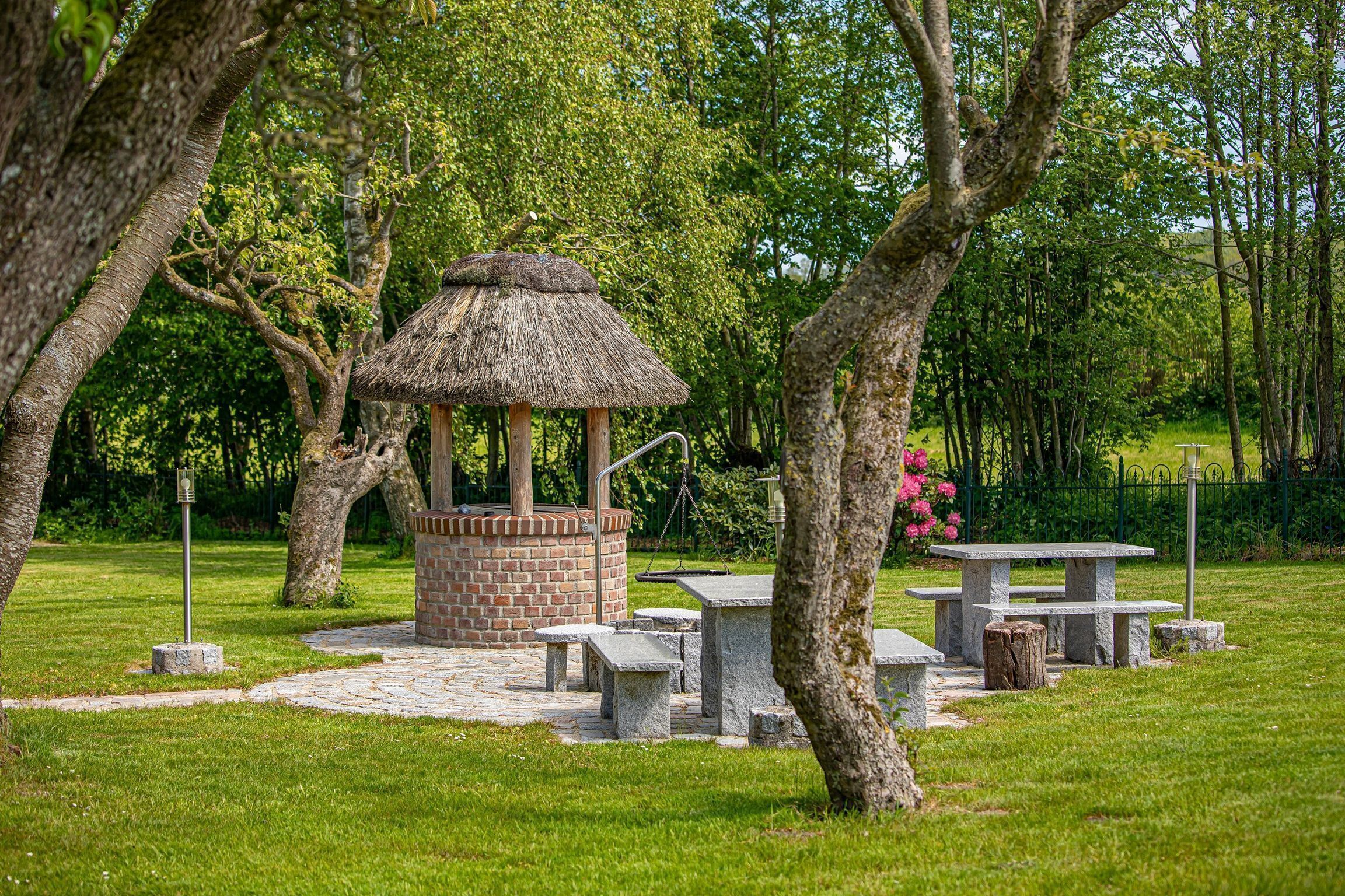 Un kiosque doté d’un toit de chaume et d’une base en briques, entouré de bancs en pierre et de tables de pique-nique, situé dans un parc.