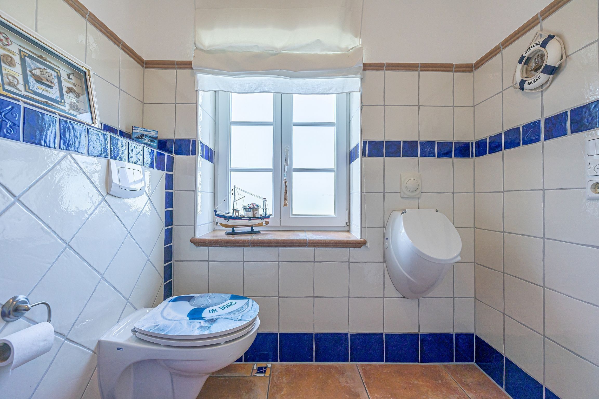 Une salle de bain équipée de toilettes, d’un urinoir et d’une fenêtre décorée avec un motif de bateau.