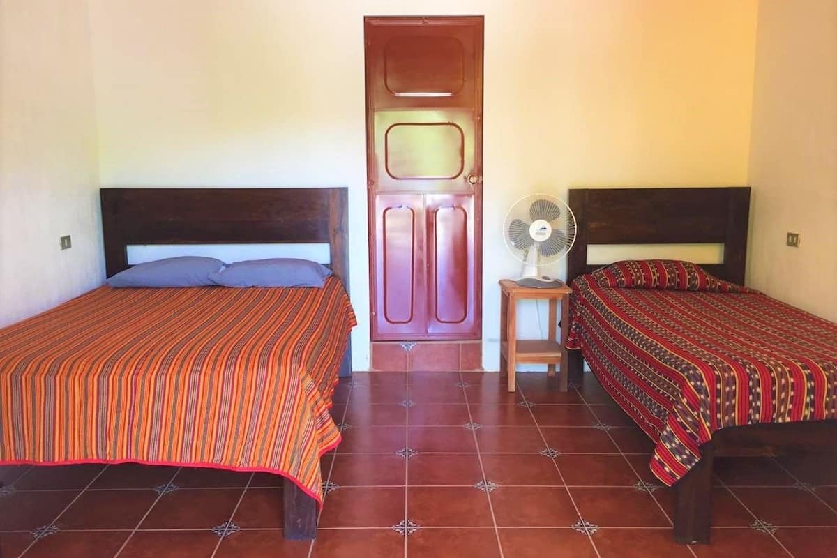 A room with two beds, a wooden door, and a fan on a small table.