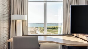 Quarto de hotel com uma janela ampla que oferece vista para a paisagem costeira, uma escrivaninha com abajur e uma televisão.