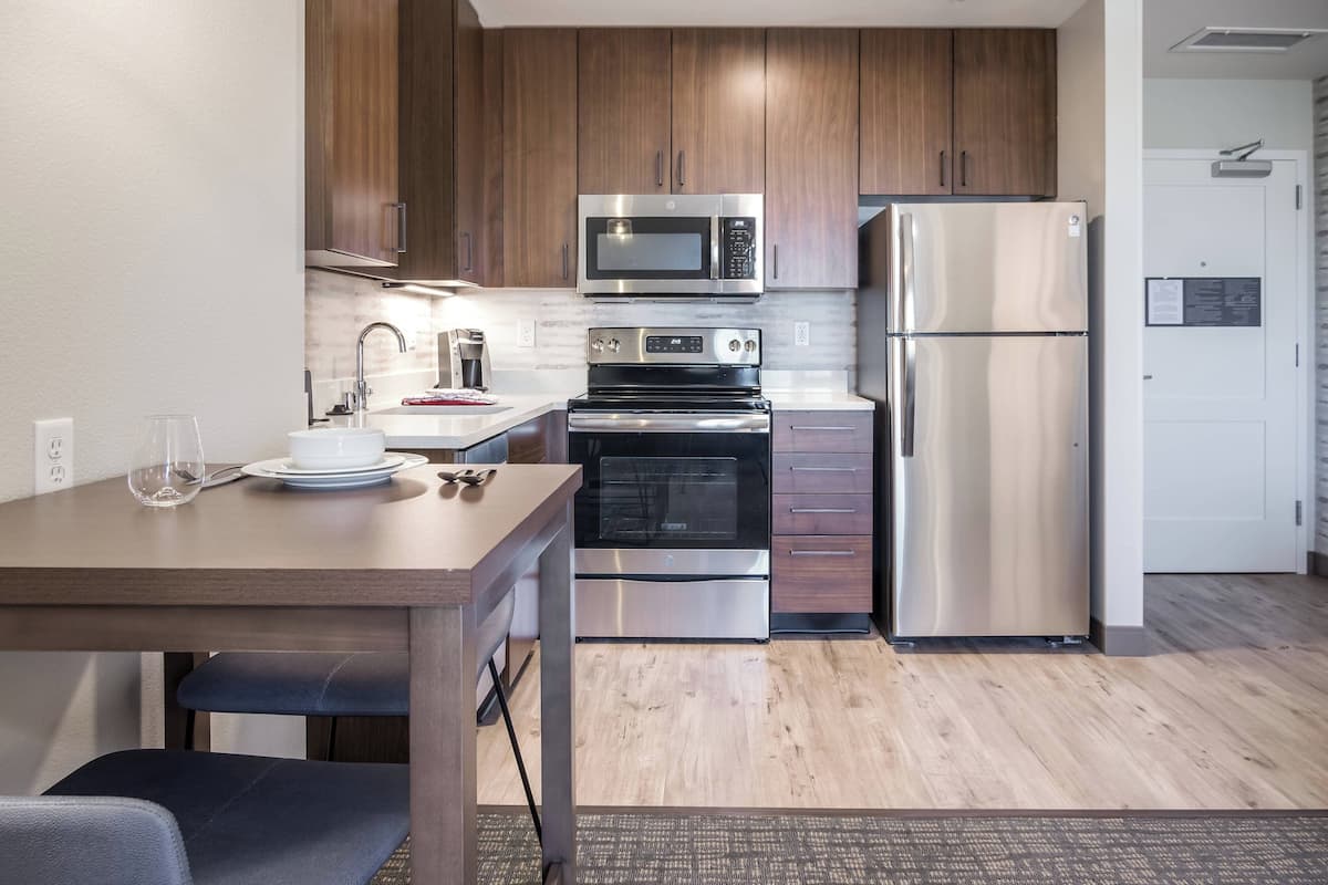 A compact kitchen with wooden cabinets, stainless steel appliances including a microwave and refrigerator, a dining table with chairs, and a small countertop with a sink.