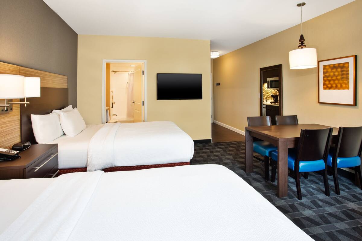 A hotel room with a large bed, a desk with chairs, a flat-screen TV, and a door leading to a bathroom.