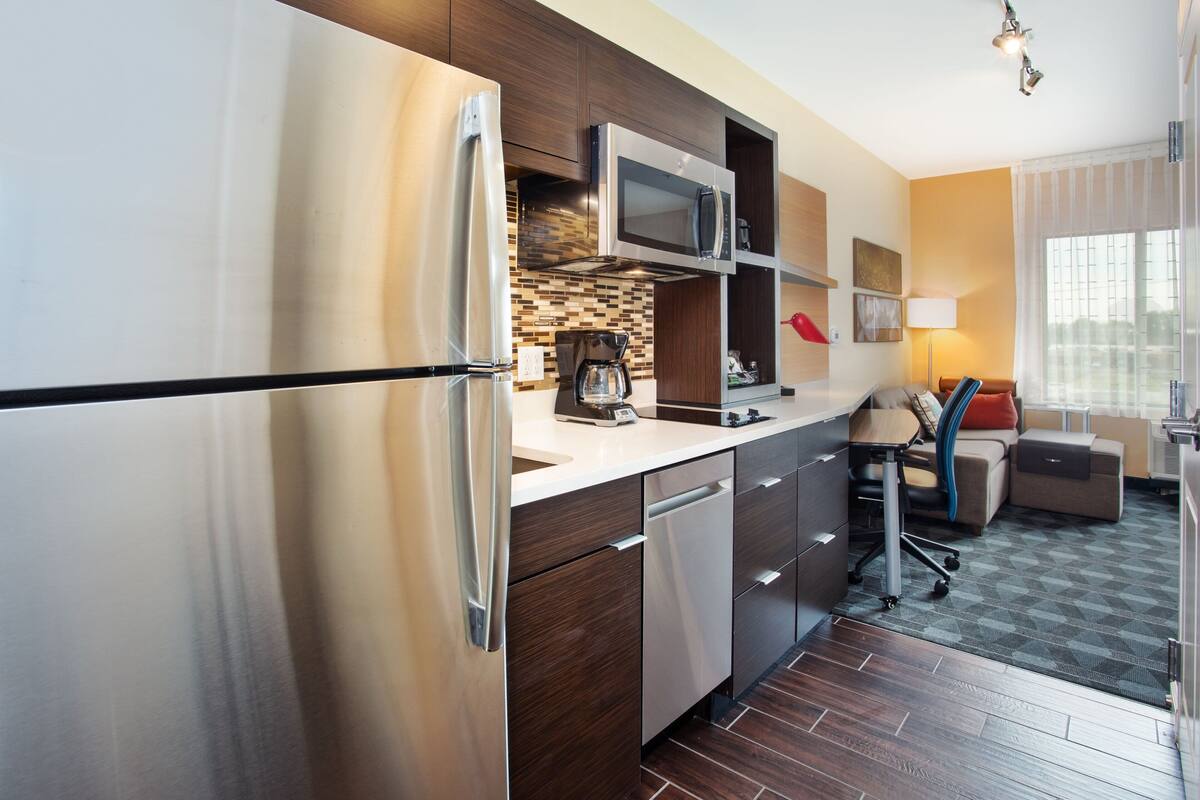 A modern kitchen with a stainless steel refrigerator, microwave, and coffee maker. There is a dining area with a table and chairs, and a sofa. The room has wooden flooring and a window with curtains.