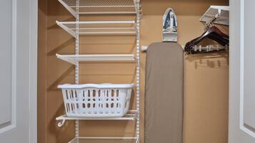 A closet with white metal shelves, a white laundry basket, and a brown ironing board.