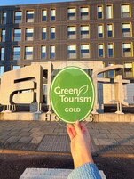 Uma mão segura uma placa verde com o texto "Green Tourism GOLD" em frente a um edifício com várias janelas e design moderno.